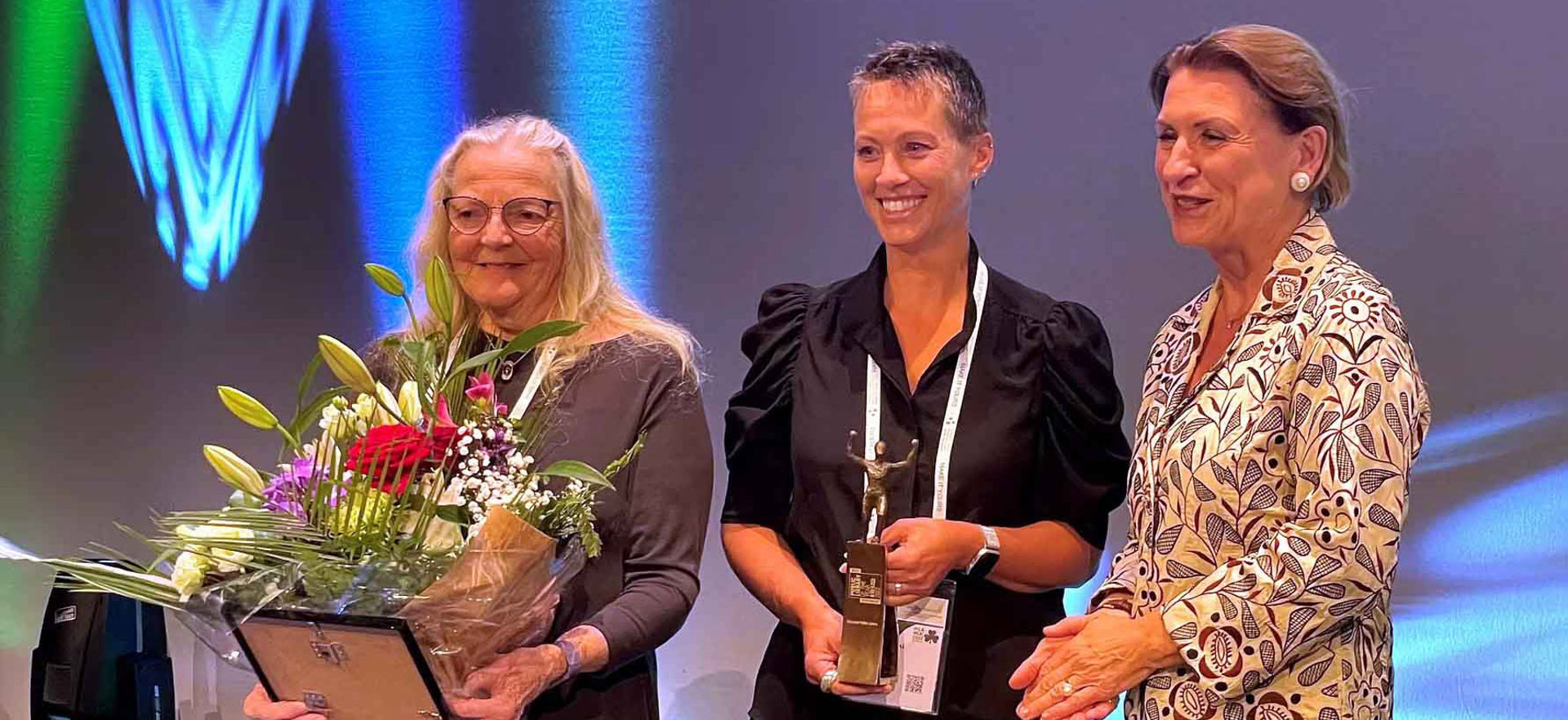 Three people smile at the camera. Two of the people hold awards and flowers.